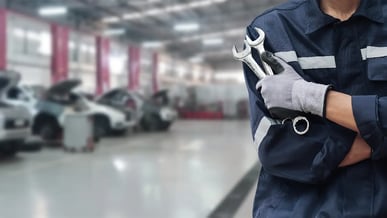 Close em um mecânico vestindo uniforme e segurando ferramentas, com oficina ao fundo