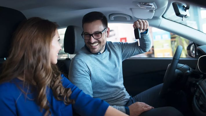 Casal dentro de carro zero, com homem no banco do motorista sorrindo e mostrando a chave para mulher