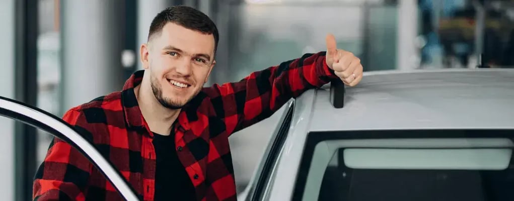 Homem sorridente com polegar para cima ao lado de carro alugado
