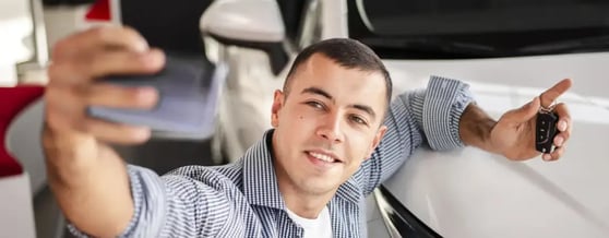 Homem sorrindo tirando uma selfie na frente de um carro branco segurando a chave na mão