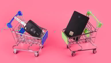 Dois carrinhos de supermercado em miniatura: um com chave de carro e outro com cartão de crédito, sobre fundo rosa