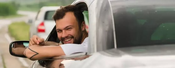 Homem sorrindo dentro do carro, apoiado na janela, visto de fora em ângulo traseiro