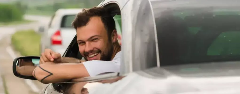 Homem sorrindo dentro do carro, apoiado na janela, visto de fora em ângulo traseiro