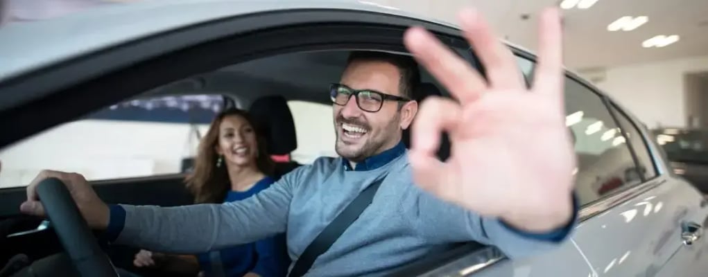 Casal feliz dentro de um carro, com homem fazendo sinal de ok para a câmera