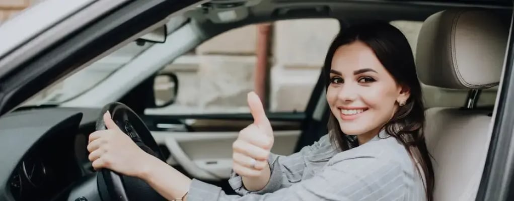 Mulher dentro de carro segurando o volante e olhando para a câmera, fazendo um sinal com o polegar para cima