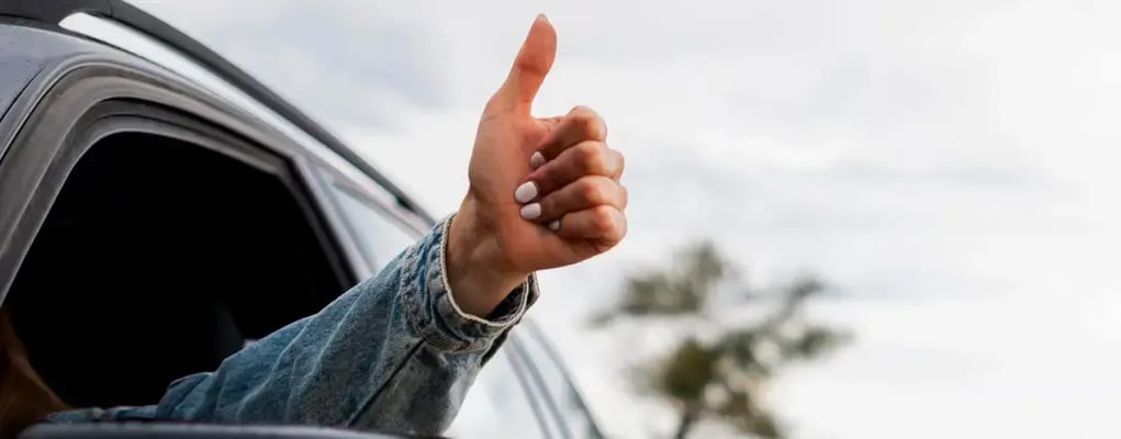 Mulher com o braço para fora da janela do carro fazendo sinal com polegar para cima