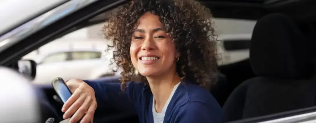 Mulher motorista de aplicativo apoiada no volante sorrindo olhando para fora da janela