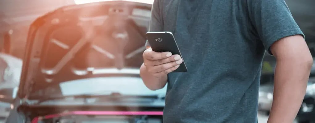 Homem mexendo no celular em frente a um carro com o capô aberto