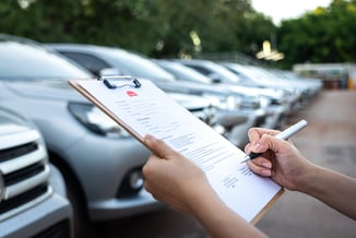 Mão preenchendo papel com logo Kovi em uma prancheta, com estacionamento de carros ao fundo