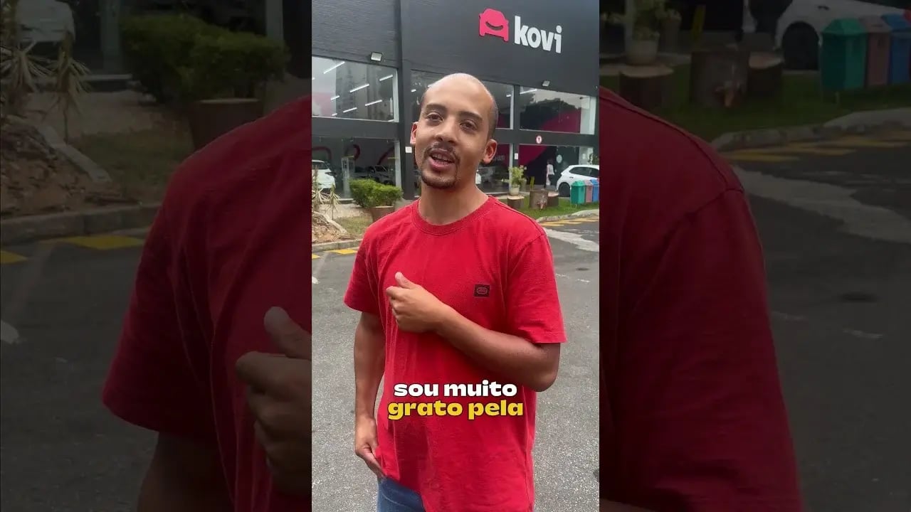 Homem de camiseta vermelha em frente a um prédio com o logo da Kovi na fachada.
